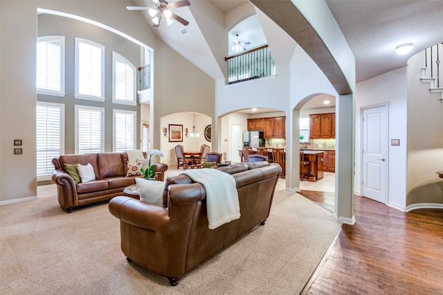 living room with ceiling fan, a stone fireplace, light carpet, and high vaulted ceiling