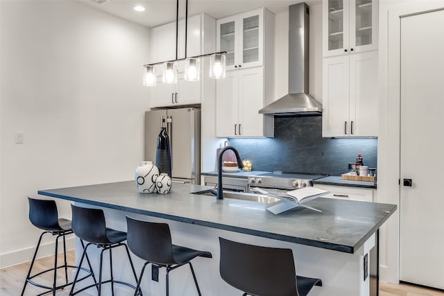 kitchen featuring white cabinets, an island with sink, hanging light fixtures, wall chimney range hood, and appliances with stainless steel finishes