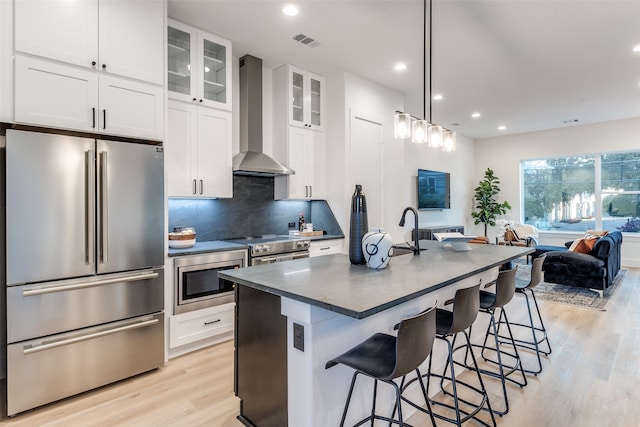 kitchen featuring an island with sink, wall chimney range hood, white cabinetry, appliances with stainless steel finishes, and a breakfast bar area