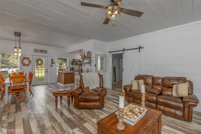 living room featuring wood ceiling, light hardwood / wood-style floors, ceiling fan with notable chandelier, a barn door, and wooden walls