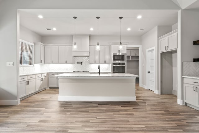 kitchen with decorative light fixtures, light wood-type flooring, a kitchen island with sink, and stainless steel appliances