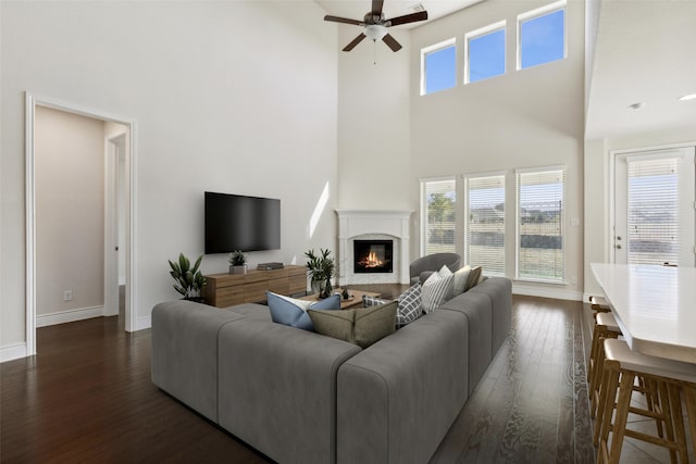 living room with a towering ceiling, ceiling fan, a large fireplace, and dark wood-type flooring