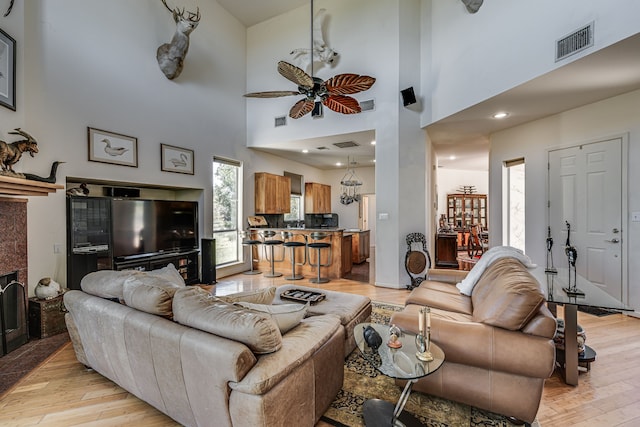 living room featuring ceiling fan, a tiled fireplace, a towering ceiling, and light hardwood / wood-style flooring