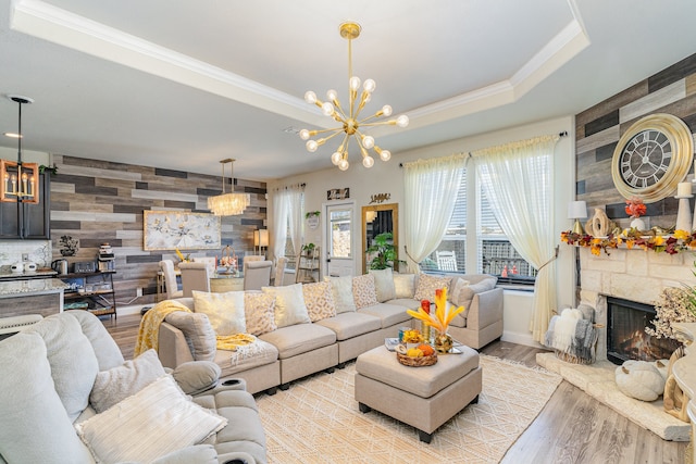 living room featuring a raised ceiling, a stone fireplace, an inviting chandelier, light hardwood / wood-style flooring, and wooden walls