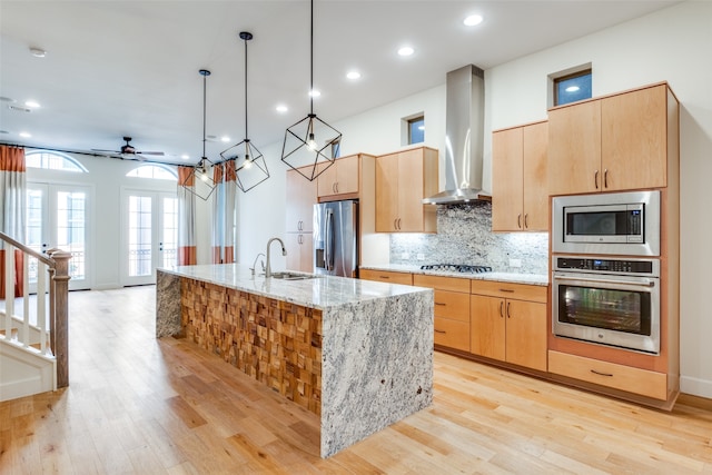 kitchen featuring decorative light fixtures, a kitchen island with sink, light stone countertops, appliances with stainless steel finishes, and wall chimney exhaust hood