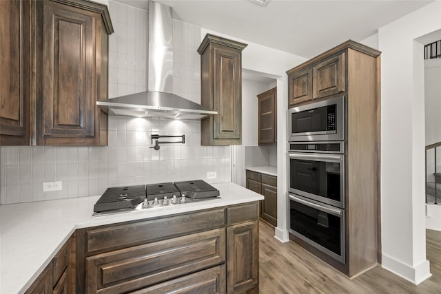 kitchen featuring dark brown cabinetry, stainless steel appliances, tasteful backsplash, wall chimney range hood, and light hardwood / wood-style flooring