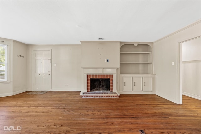 unfurnished living room with a brick fireplace, built in shelves, wood-type flooring, and ornamental molding