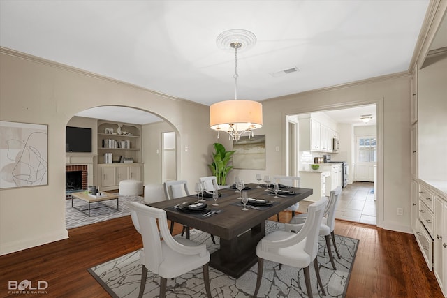 dining space featuring dark wood-type flooring, ornamental molding, built in shelves, and a brick fireplace