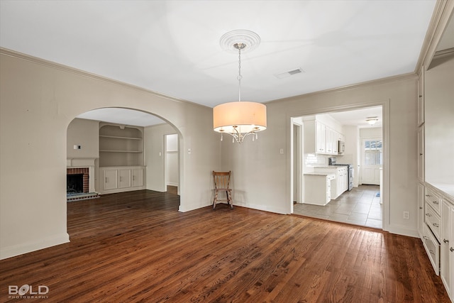 unfurnished dining area featuring dark wood-type flooring, ornamental molding, built in shelves, and a brick fireplace