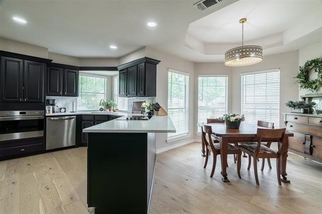 kitchen with a raised ceiling, hanging light fixtures, kitchen peninsula, appliances with stainless steel finishes, and light wood-type flooring