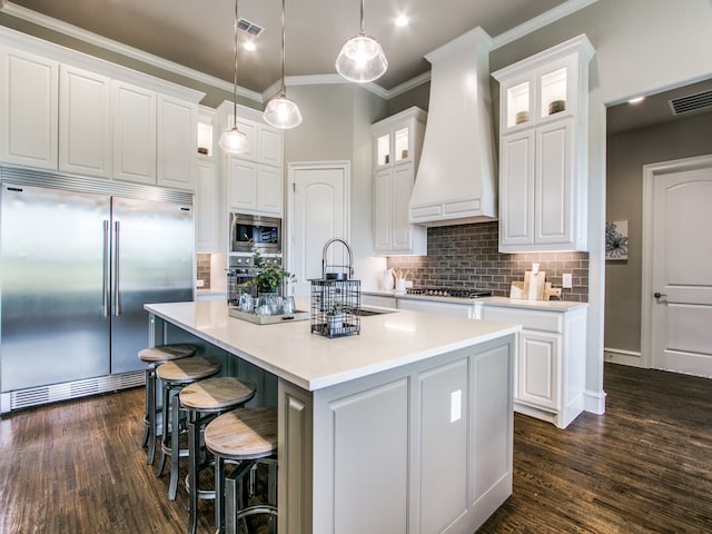 kitchen featuring premium range hood, an island with sink, white cabinetry, built in appliances, and dark hardwood / wood-style flooring