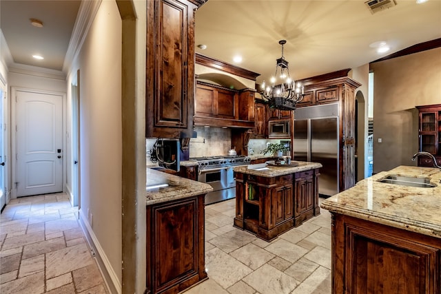 kitchen with built in appliances, crown molding, light stone countertops, sink, and an island with sink