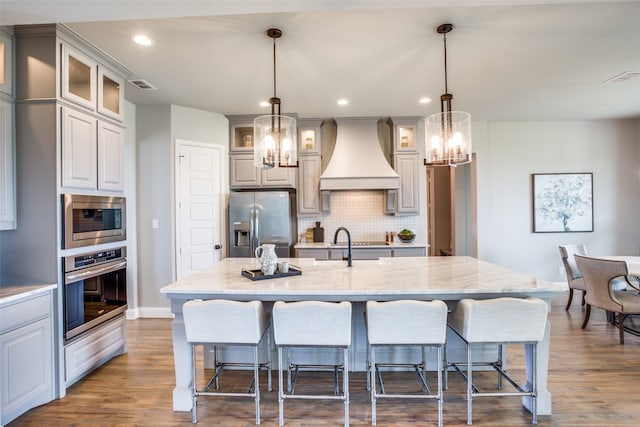 kitchen with hanging light fixtures, a large island with sink, dark wood-type flooring, appliances with stainless steel finishes, and custom range hood