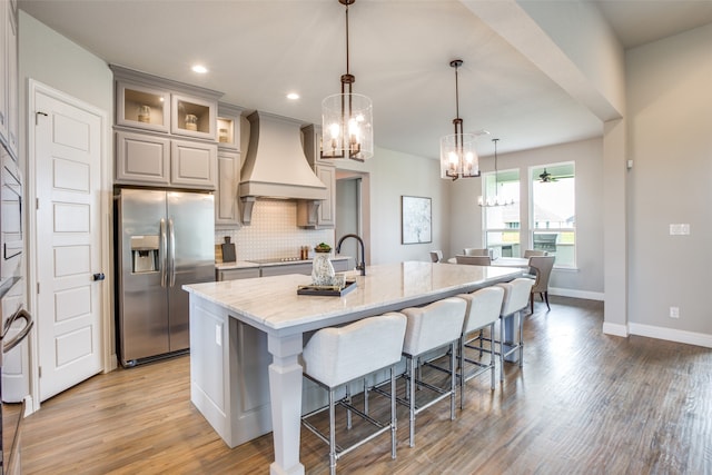 kitchen with custom exhaust hood, a kitchen island with sink, light hardwood / wood-style flooring, stainless steel refrigerator with ice dispenser, and light stone countertops