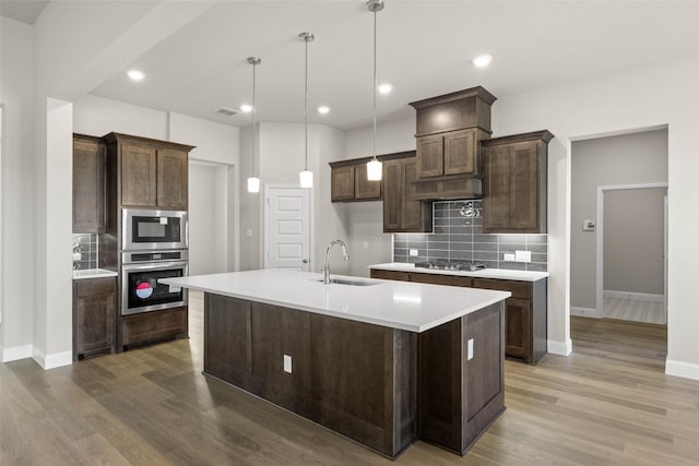 kitchen with a center island with sink, stainless steel appliances, tasteful backsplash, a sink, and dark brown cabinets