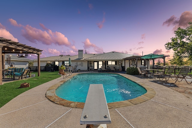 pool at dusk featuring a diving board, a pergola, a patio area, and a lawn