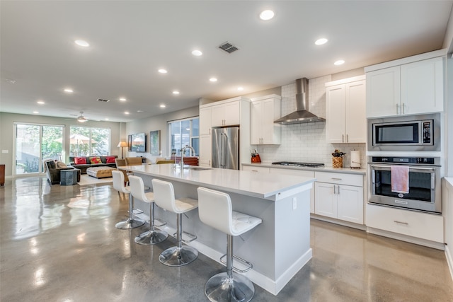 kitchen with an island with sink, wall chimney exhaust hood, white cabinetry, appliances with stainless steel finishes, and a breakfast bar