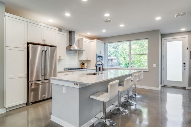 kitchen with white cabinets, a center island with sink, appliances with stainless steel finishes, and wall chimney range hood