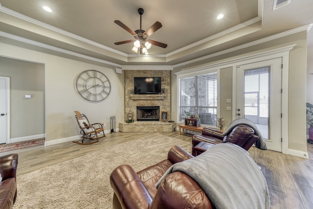 living room featuring crown molding, a stone fireplace, a tray ceiling, and light hardwood / wood-style flooring