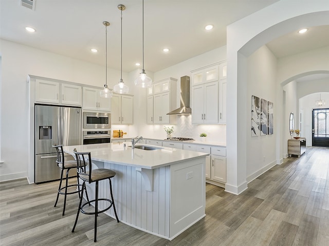 kitchen featuring a center island with sink, appliances with stainless steel finishes, hanging light fixtures, wall chimney exhaust hood, and white cabinets