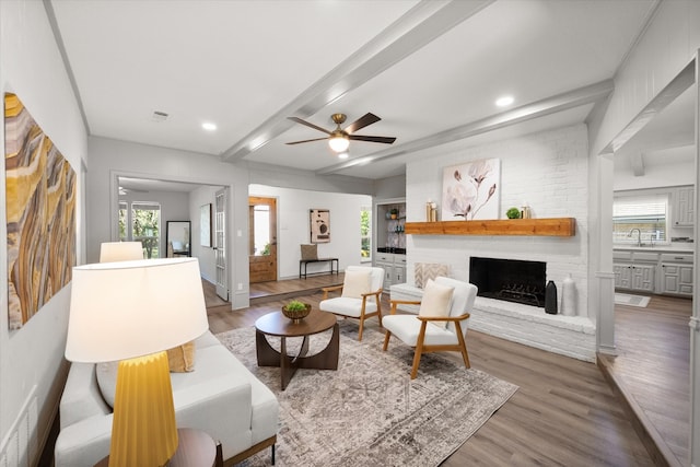 living room featuring a brick fireplace, beam ceiling, ceiling fan, hardwood / wood-style flooring, and sink