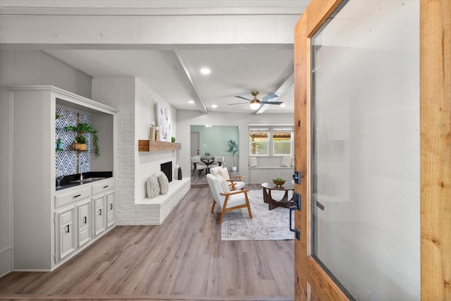 living room with a brick fireplace, light hardwood / wood-style floors, beamed ceiling, and ceiling fan