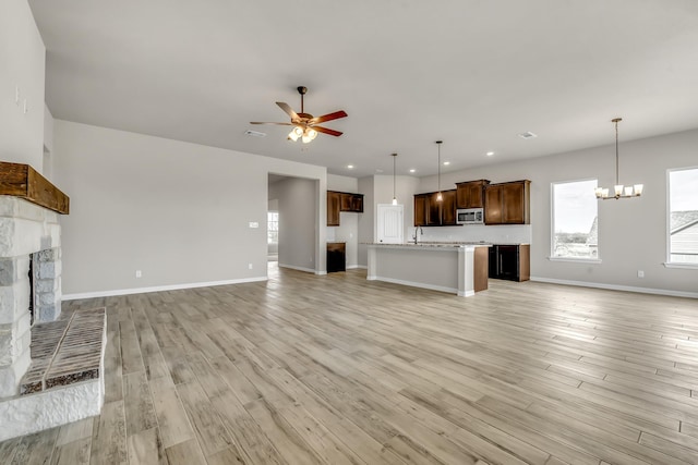 unfurnished living room with sink, ceiling fan with notable chandelier, a stone fireplace, and light hardwood / wood-style floors