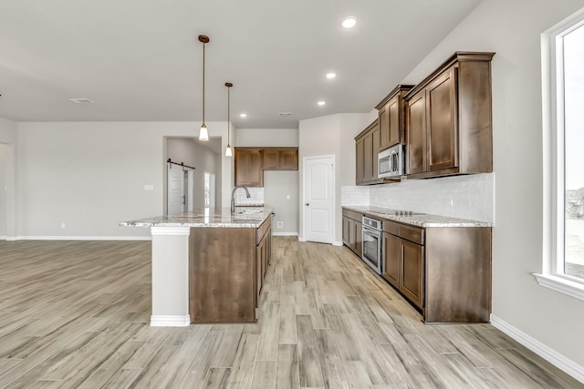 kitchen with pendant lighting, stainless steel appliances, a kitchen island with sink, light stone counters, and a barn door