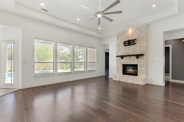 unfurnished living room with ceiling fan, a tray ceiling, a stone fireplace, and dark hardwood / wood-style floors