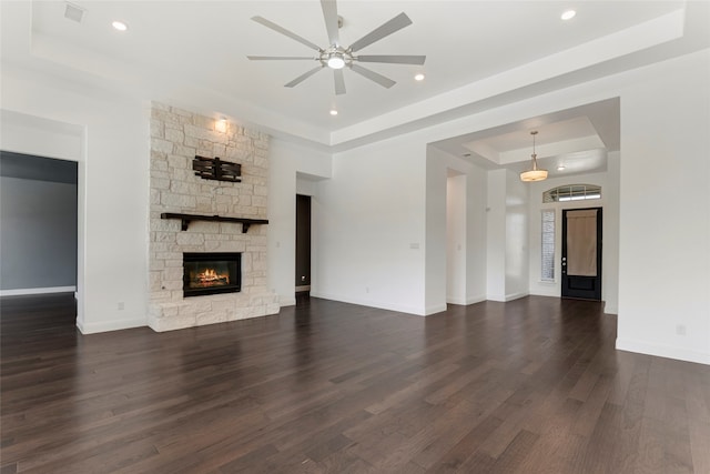 unfurnished living room with ceiling fan, a stone fireplace, a raised ceiling, and dark hardwood / wood-style floors