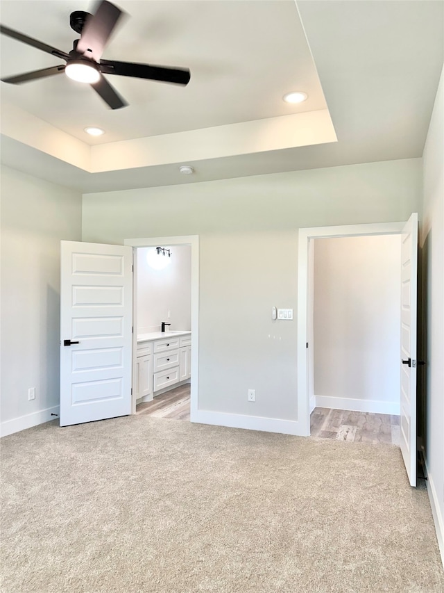 unfurnished bedroom with a tray ceiling, ensuite bath, ceiling fan, and light colored carpet