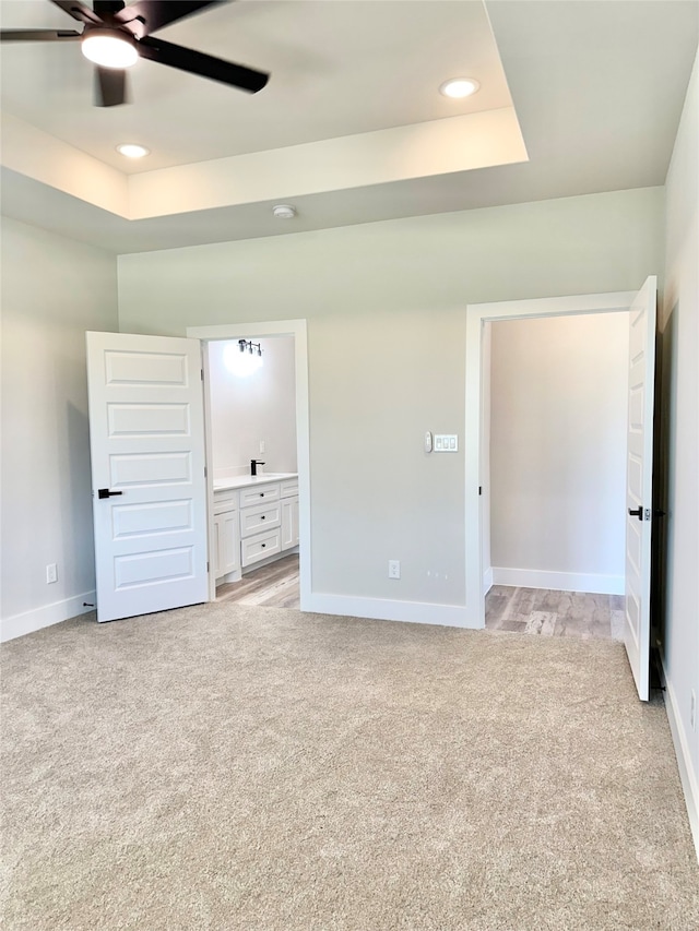 unfurnished bedroom featuring a raised ceiling, ensuite bath, ceiling fan, and light colored carpet