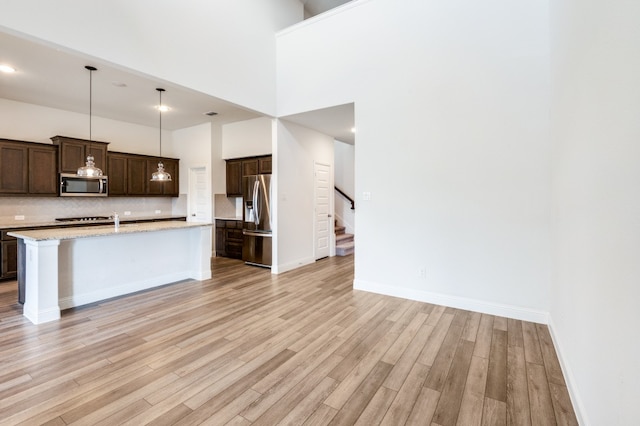 kitchen featuring appliances with stainless steel finishes, hanging light fixtures, a towering ceiling, light hardwood / wood-style flooring, and a kitchen island with sink