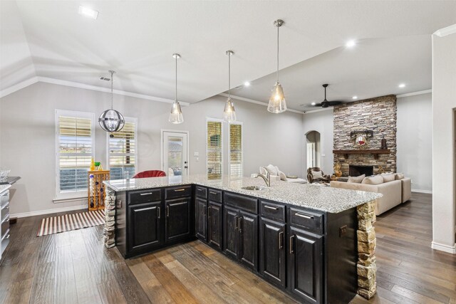 kitchen with sink, a stone fireplace, light stone counters, an island with sink, and ceiling fan with notable chandelier