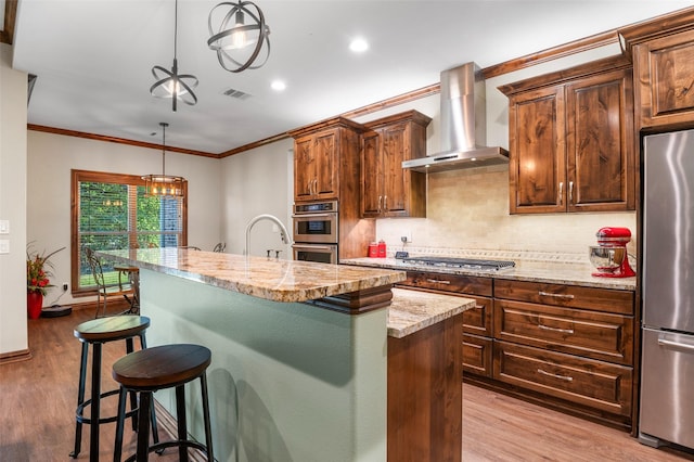 kitchen featuring wall chimney exhaust hood, stainless steel appliances, light hardwood / wood-style flooring, an island with sink, and pendant lighting