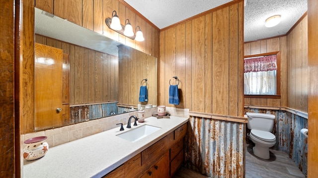 bathroom with wooden walls, toilet, vanity, ornamental molding, and a textured ceiling