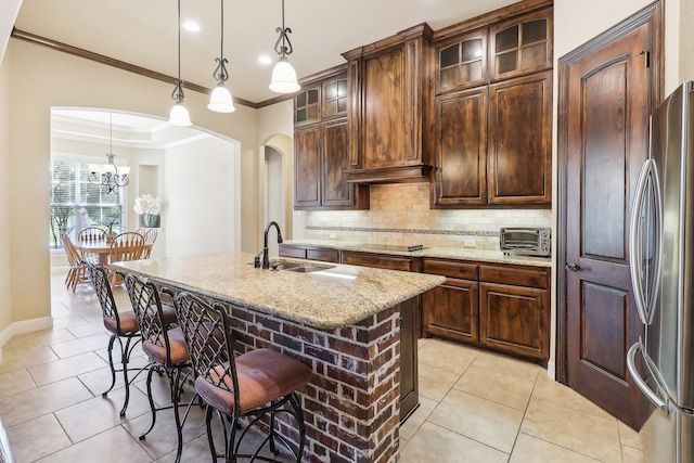 kitchen with dark brown cabinets, a kitchen island with sink, decorative light fixtures, light stone countertops, and stainless steel fridge