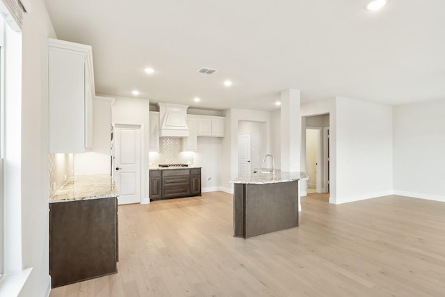 kitchen featuring backsplash, a center island with sink, custom range hood, dark brown cabinets, and white cabinetry