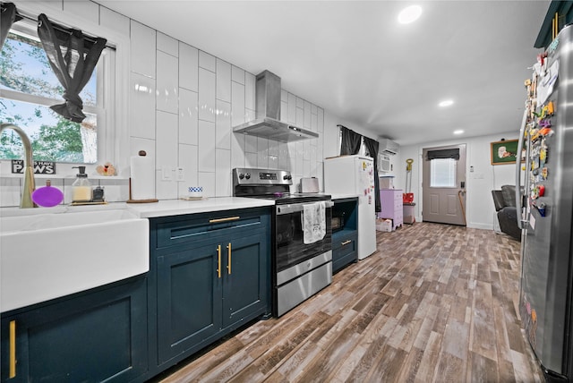 kitchen with wall chimney exhaust hood, light hardwood / wood-style flooring, backsplash, appliances with stainless steel finishes, and blue cabinetry