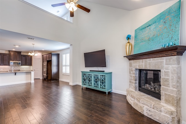 unfurnished living room featuring ceiling fan with notable chandelier, a towering ceiling, sink, a stone fireplace, and dark hardwood / wood-style floors