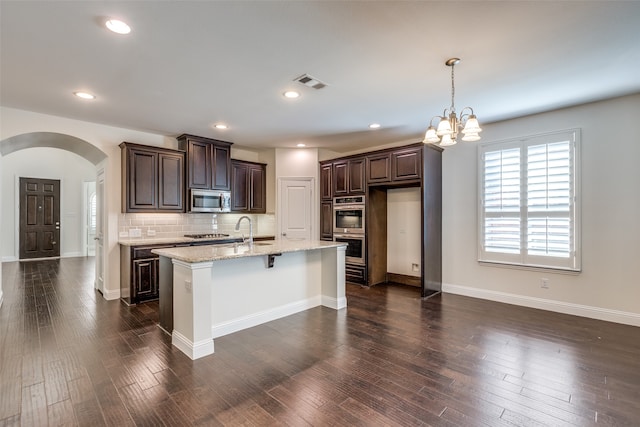 kitchen with light stone countertops, dark wood-type flooring, a kitchen island with sink, and stainless steel appliances