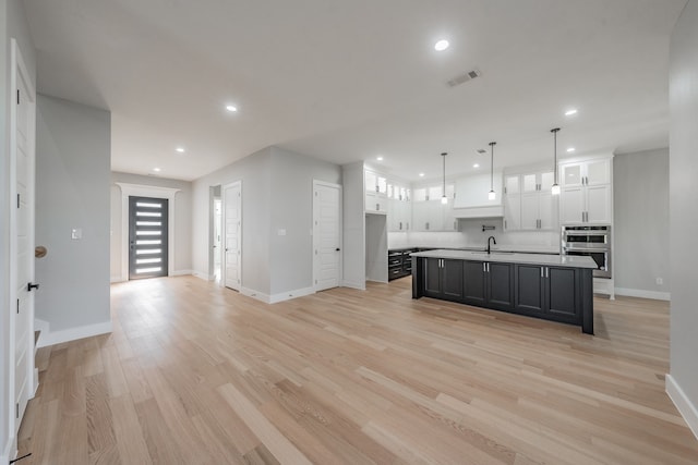 kitchen featuring an island with sink, white cabinetry, light wood-type flooring, double oven, and decorative light fixtures