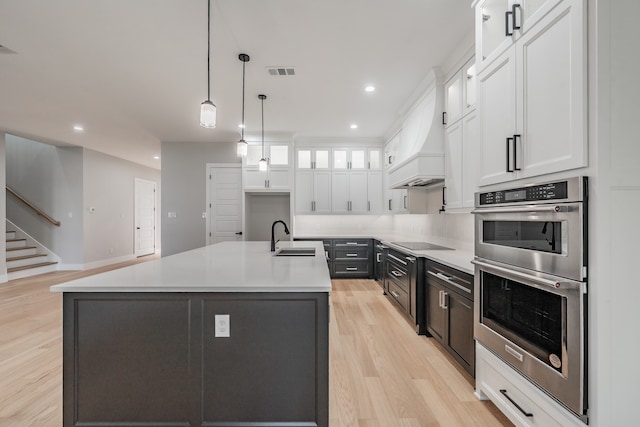 kitchen featuring an island with sink, stainless steel double oven, white cabinetry, hanging light fixtures, and light hardwood / wood-style flooring