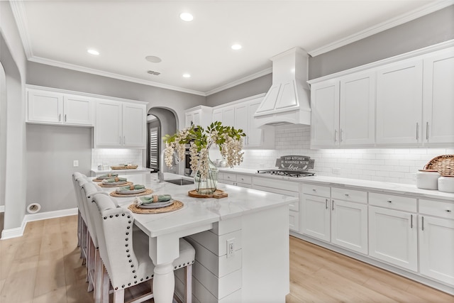 kitchen featuring light wood-type flooring, premium range hood, a center island with sink, and white cabinetry