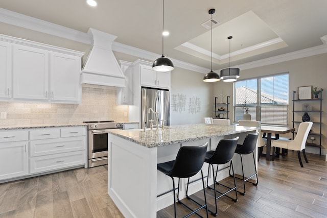 kitchen featuring custom exhaust hood, an island with sink, white cabinetry, light wood-type flooring, and appliances with stainless steel finishes