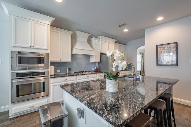 kitchen with dark wood-type flooring, a center island with sink, appliances with stainless steel finishes, and custom range hood