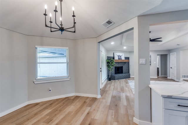 unfurnished dining area with ceiling fan with notable chandelier, light wood-type flooring, and a brick fireplace