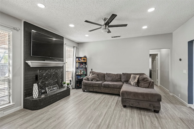 living room with a brick fireplace, light hardwood / wood-style floors, a textured ceiling, and ceiling fan