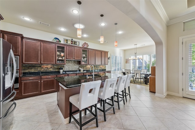 kitchen featuring ornamental molding, a center island with sink, dark stone countertops, stainless steel fridge with ice dispenser, and hanging light fixtures