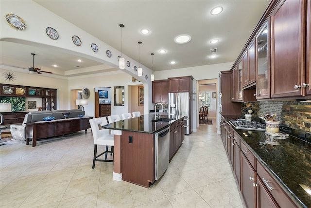 kitchen with stainless steel appliances, ceiling fan, pendant lighting, dark stone countertops, and an island with sink